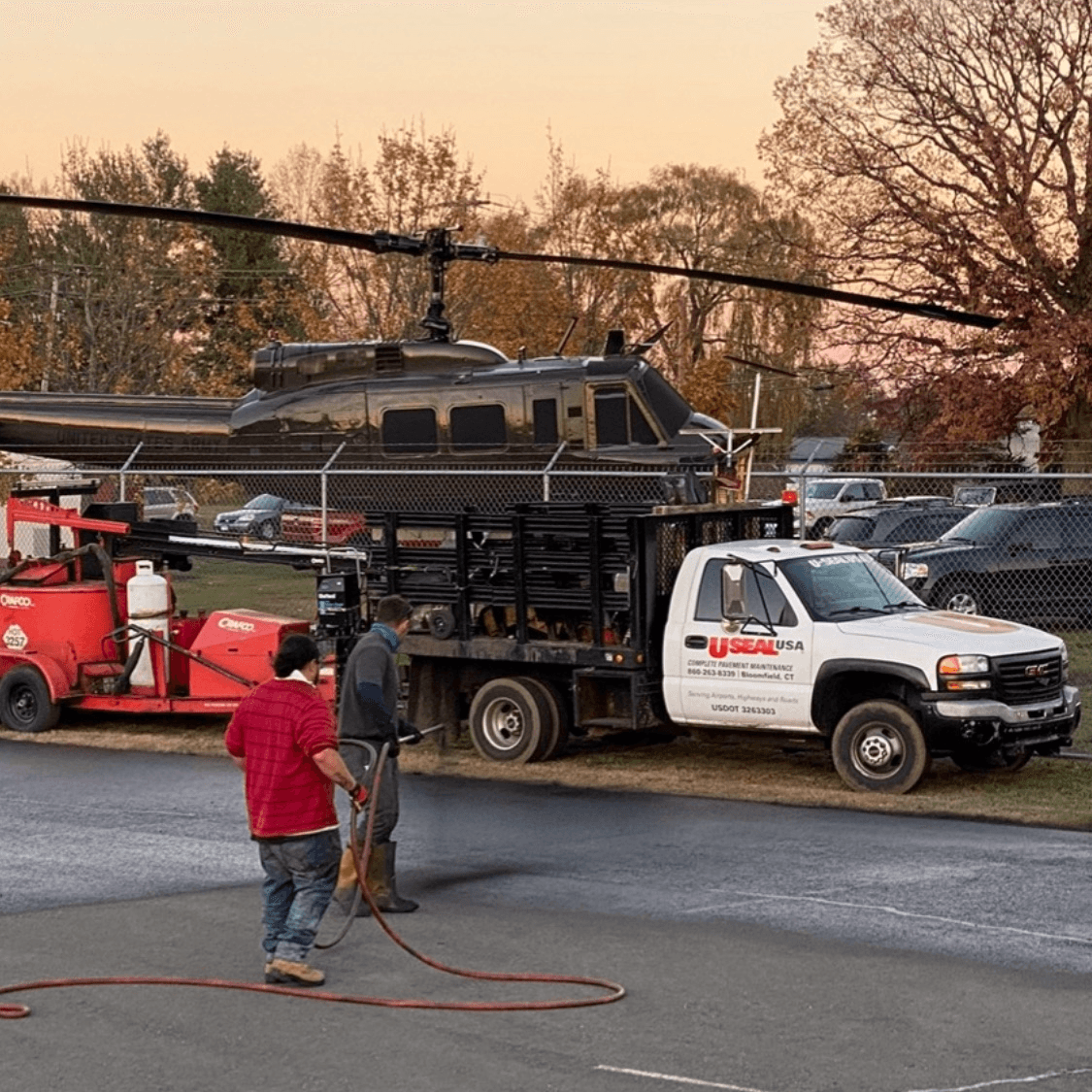 U-SEAL USA crew with branded service truck at a Connecticut helicopter memorial, freshly sealed asphalt in foreground at sunset